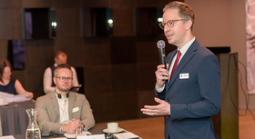 BMBF-Referent Alexander Hochradel hält eine Rede bei der Verleiung des Berufsbildungspreis Lettland 2024. Im Hintergrund sind verschwommen drei sitzende Zuhörer zu sehen.