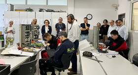 People (the Costa Rican delegation accompanied) in a training room with electrical engineering, sitting and standing, listening to explanations