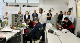 Menschen (die costa-ricanische Delegation mit Begleitung) in Ausbildungsraum mit Elektrotechnik, sitzend und stehend, hören Erläuterungen zu