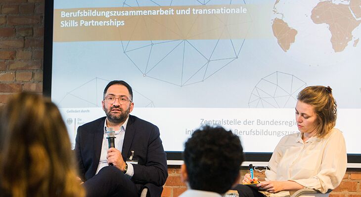 Ein Mann mit Mikrofon und eine Frau, die sich Notizen macht, sitzen auf einem Posium bei einer Konferenz, im Hintergrund eine Folie mit der Aufschrift "Berufsbildungszusammenarbeit und transnationale Skills Partnerships