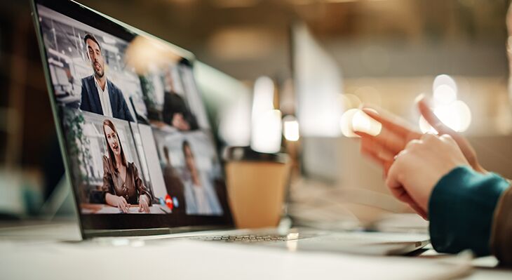 Laptop auf Schreibtisch. Der Bildschirm zeigt eine Videokonferenz. Im rechten Rand verschwommene Hände.